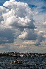 A small boat sails alone on the sea, with Istanbul's historic mosques and the city skyline in the background. Heavy clouds and dramatic light in the sky.