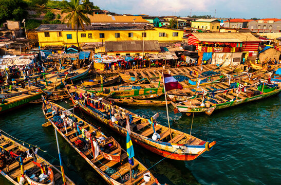 View of colorful fishing boats bobbing gently in the harbour waters under a bright sky, with vibrant buildings lining the shore, Elmina, Central Region, Ghana.