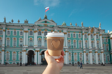 paper cup of drink with carved heart in woman's palm on background of Winter palace l in St....