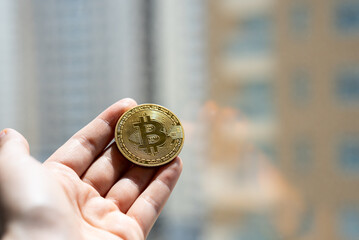 Close-up of a person holding golden Bitcoin cryptocurrency coin against blurred city background,...