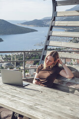 Digital nomad working remotely on laptop at seaside terrace; coastal town and islands behind....