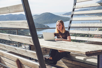 Digital nomad working remotely on laptop at seaside terrace; coastal town and islands behind....