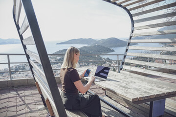 Digital nomad working remotely on laptop at seaside terrace; coastal town and islands behind....