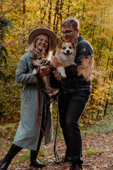 A couple is happily holding two corgi dogs in the woods