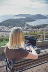 Digital nomad working remotely on laptop at seaside terrace; coastal town and islands behind....