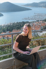 Digital nomad working remotely on laptop at seaside terrace; coastal town and islands behind....