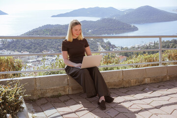 Digital nomad working remotely on laptop at seaside terrace; coastal town and islands behind....