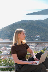 Digital nomad working remotely on laptop at seaside terrace; coastal town and islands behind....