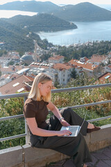 Digital nomad working remotely on laptop at seaside terrace; coastal town and islands behind....