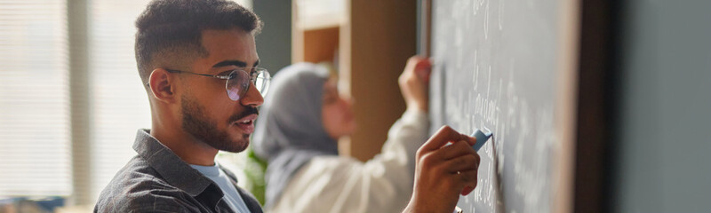 Website header shot of young adult Black man writing on chalkboard in language school classroom, while young adult Middle Eastern woman standing in background also writing on board © Seventyfour