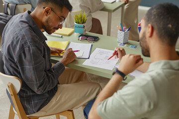 Two young adult men, one Black and one Middle Eastern, sitting at desk studying language materials...