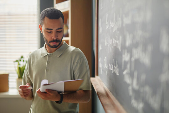 Young adult Middle Eastern man standing in classroom holding notebook, reading or preparing lesson in front of chalkboard with handwritten text, language school educational setting