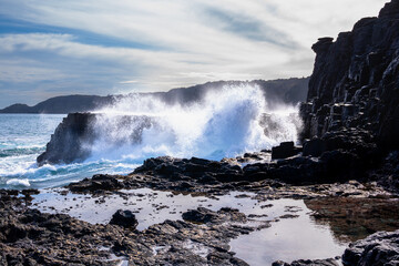 Huge ocean wave crashes against rugged volcanic cliffs, creating dramatic spray under a cloudy sky.
