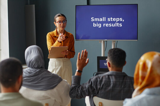 Young adult Caucasian woman standing in front of screen holding clipboard, teaching diverse group of adults in language school classroom, students raising hands to participate - Powered by Adobe