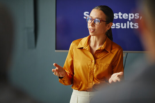 Young adult Caucasian woman standing in front of screen gesturing while explaining language lesson to students in classroom setting at language school