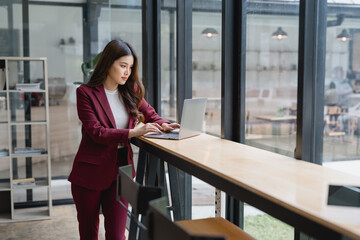 Young Asian businesswoman standing and using laptop computer near window in modern office, focused entrepreneur working on online project, female executive using technology for business tasks