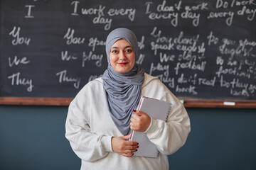 Portrait of young adult Middle Eastern woman wearing hijab standing in front of chalkboard holding notebooks smiling at camera in language school classroom setting