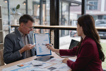 Business partners reviewing financial reports, analyzing charts and graphs using mobile phone and pen, discussing company performance and planning future strategies in a modern office environment
