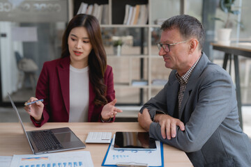 Asian businesswoman showing explaining project results to senior businessman during meeting, using laptop and financial documents in modern office