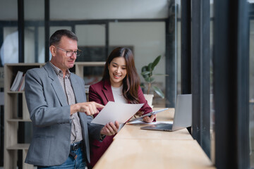 Two businesspeople are standing by a desk, examining paperwork together and using a laptop in a modern office setting, engaging in collaborative discussion and problem-solving