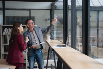 Businessman pointing outside window while discussing with businesswoman using laptop and documents in modern office, talking about future vision, planning new project, developing marketing strategy © Tj