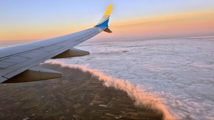 An airplane wing with a yellow and blue tip flies over clouds and cities	
