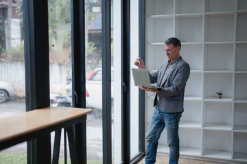 Businessman working on a laptop and jotting down notes while standing by a window in a modern office, showcasing a bright and professional workspace filled with contemporary design elements