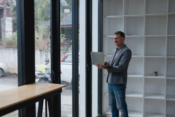 Fototapeta premium Businessman wearing glasses and a grey jacket, standing and working on his laptop in a modern office space filled with natural light, smiling happily while engaging in his tasks
