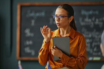 Caucasian young adult woman standing in classroom holding digital tablet, gesturing while explaining lesson content, blackboard with handwritten notes visible in background, language school setting