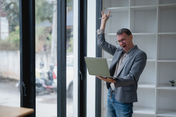 Mature businessman celebrating a successful online deal while holding a laptop and making a rock and roll hand gesture in a modern office, radiating joy and enthusiasm