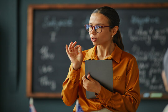 Caucasian young adult woman standing in classroom holding digital tablet, gesturing while explaining lesson content, blackboard with handwritten notes visible in background, language school setting