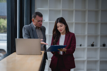 Two businesspeople wearing suits are discussing and working together in a modern office using laptop and clipboard, analyzing financial data and planning new strategy