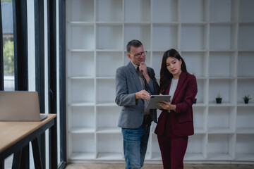 Fototapeta premium Businessman and businesswoman discussing over digital tablet in modern office, they're standing next to a desk with a laptop and a white bookcase