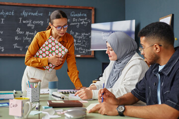 Young adult Caucasian woman standing and assisting Middle Eastern woman in hijab and young adult Black man sitting at desk, studying language school materials together