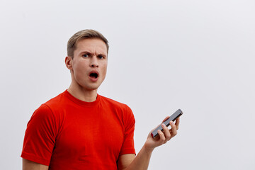 Confused man in bright red t-shirt holding smartphone with surprised facial expression isolated on...