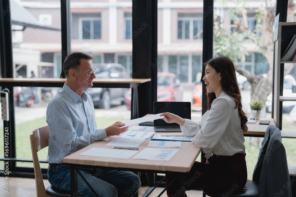 Poster two business partners are exchanging documents with financial charts and graphs while sitting at a d