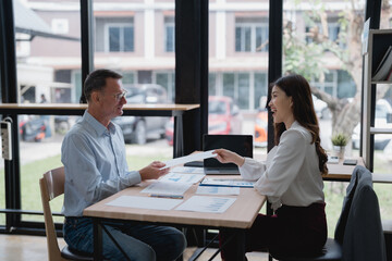 Two business partners are exchanging documents with financial charts and graphs while sitting at a desk in a modern office, working together on a project