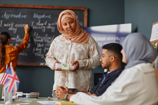 Muslim woman demonstrating sticker note with new vocabulary to diverse young adults in language school classroom, British flag and chalkboard with English phrases visible in background