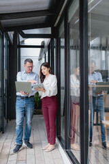Businessman and businesswoman standing in a modern office corridor, collaborating and discussing work while comparing data on a laptop and digital tablet, fostering teamwork and communication