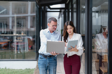 Senior businessman showing something on his laptop to young businesswoman holding tablet, discussing work while walking outside modern office building