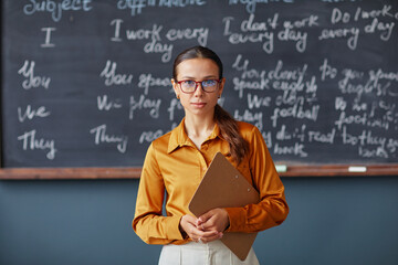 Portrait of young adult Caucasian woman wearing glasses standing in front of chalkboard holding...