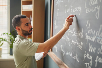 Young adult Black man writing English sentences on chalkboard in classroom holding notebook,...