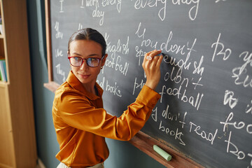 Caucasian young adult woman writing English phrases on chalkboard, wearing glasses, standing in classroom, teaching language lesson, looking over shoulder toward students