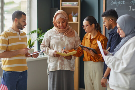 Group of diverse young adult men and women collaborating in language school classroom, holding notebooks and digital tablet, discussing lesson material and taking notes together