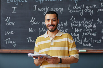 Portrait of young adult Middle Eastern man standing in front of chalkboard holding notebooks...