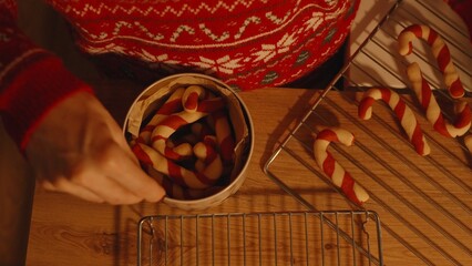 Hand Placing Candy Cane Cookies into Round Tin Beside Cooling Rack and Tabletop, Overhead Angle, Cookie Storage Concept