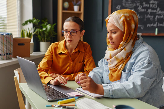 Caucasian young adult woman and Middle Eastern young adult woman wearing hijab studying together at language school classroom, using laptop and taking notes with pens and notebooks