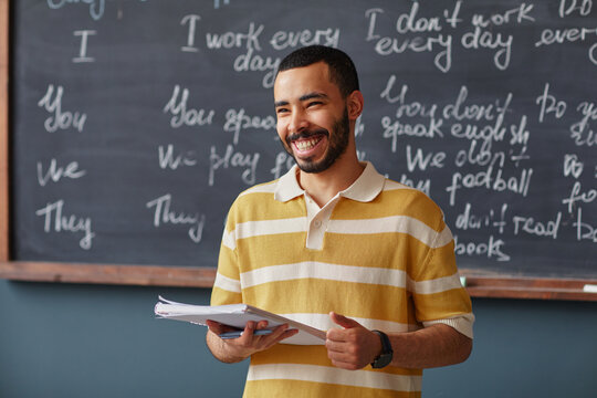 Portrait of young adult Middle Eastern man smiling while holding notebook, standing in front of chalkboard with handwritten English phrases in language school classroom setting