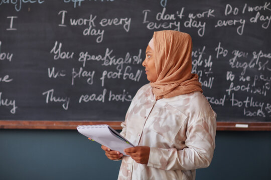 Muslim biracial woman standing in front of chalkboard holding notebook, teaching language lesson in classroom, looking away from camera, wearing headscarf