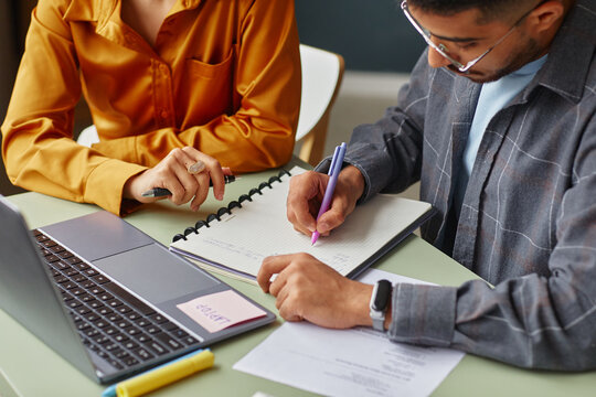 Young adult Caucasian woman and young adult South Asian man studying together at table, writing in notebook, using laptop and reviewing documents in language school classroom setting